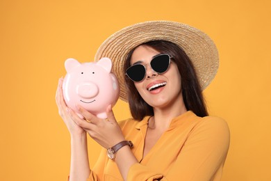 Photo of Young woman in stylish sunglasses and straw hat with piggy bank on orange background