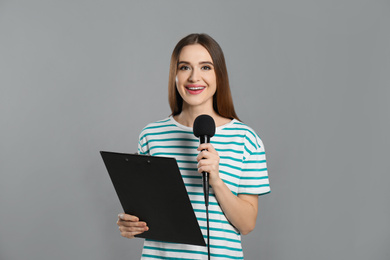 Young female journalist with microphone and clipboard on grey background