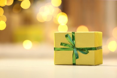 Photo of Beautifully wrapped gift box on white table against blurred festive lights, space for text. Christmas celebration
