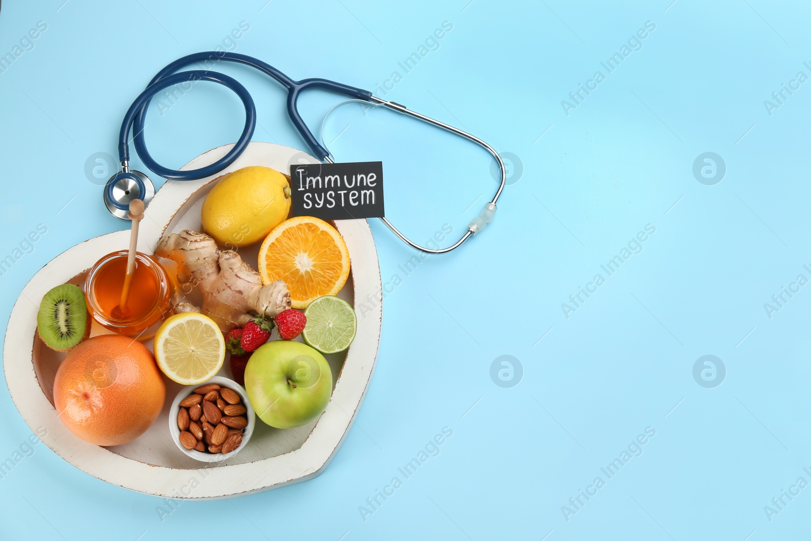 Photo of Heart shaped tray of healthy products, stethoscope and card with phrase Immune System on light blue background, flat lay. Space for text