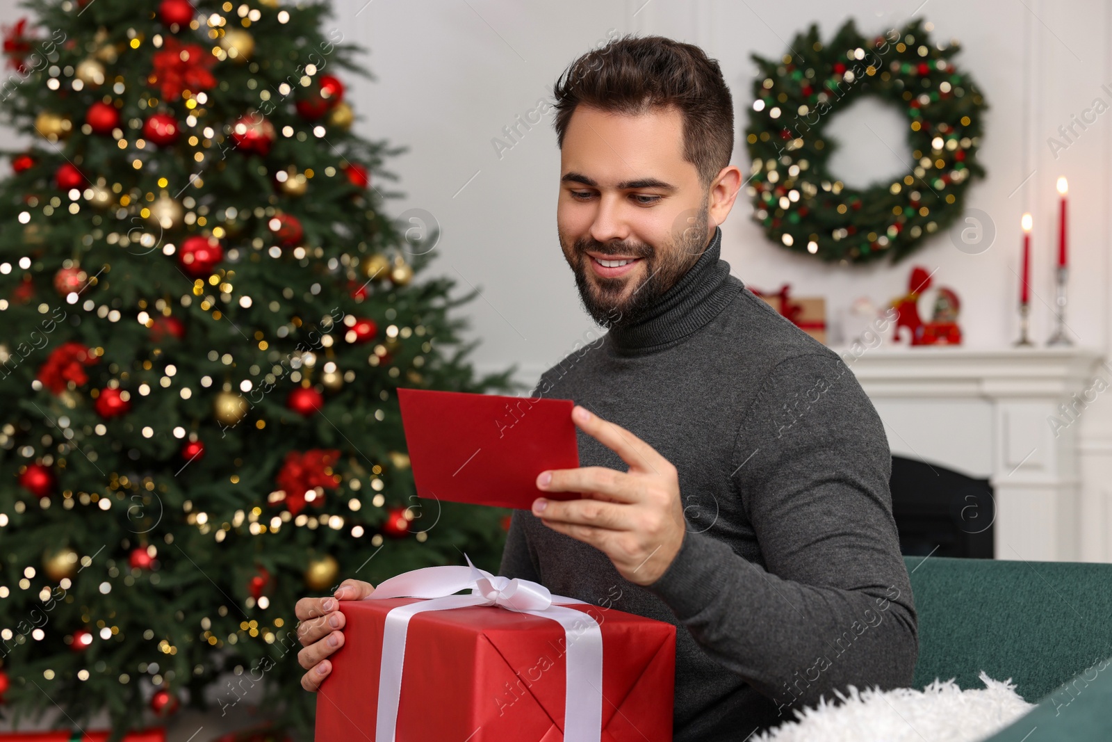 Photo of Happy young man with Christmas gift reading greeting card at home