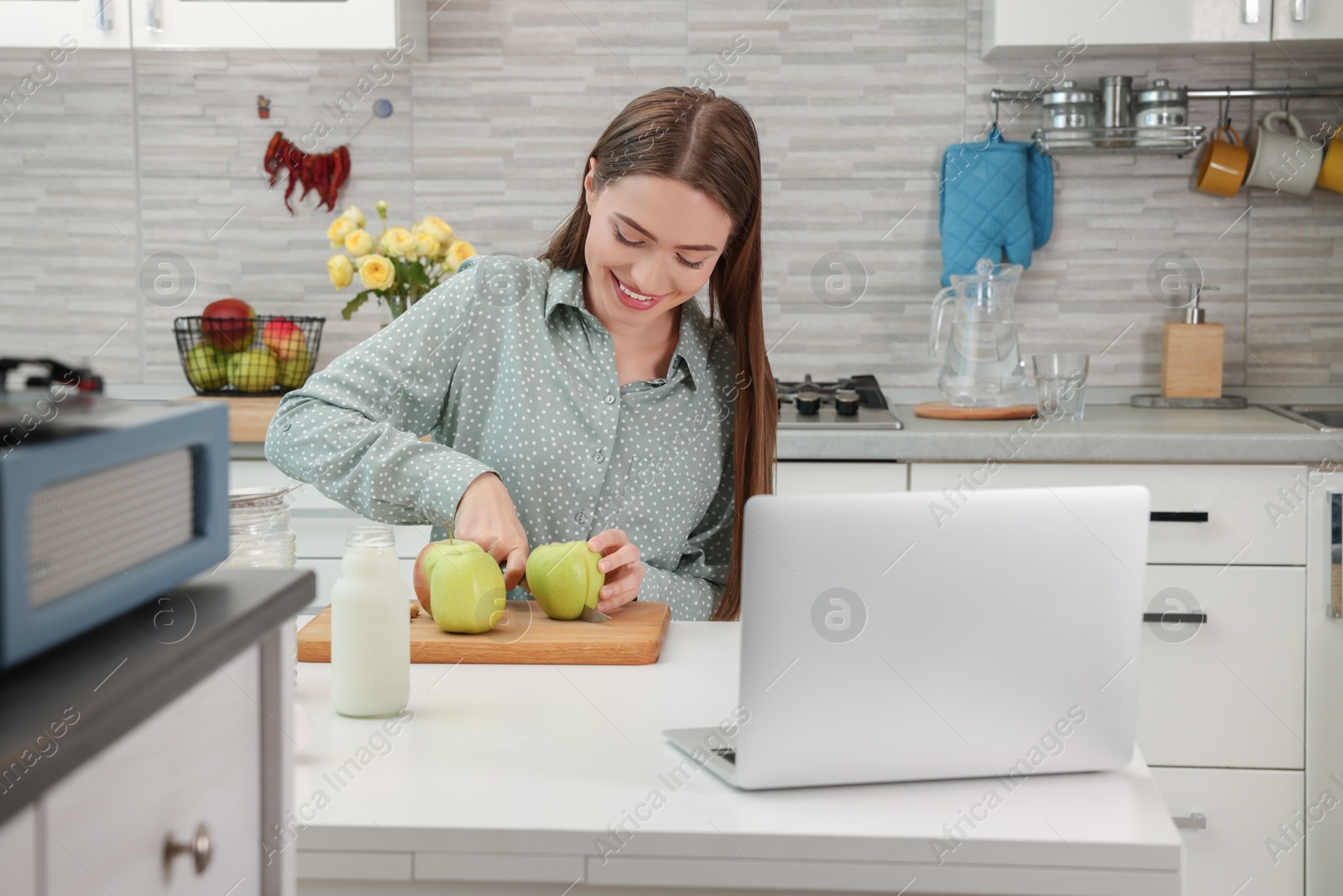 Photo of Happy young woman making apple cake while watching online cooking course via laptop in kitchen. Time for hobby