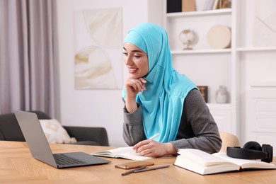 Muslim woman writing notes near laptop at wooden table in room