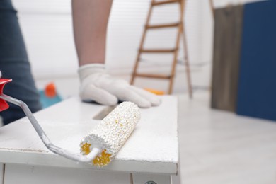 Photo of Man using roller to paint bekvam with white dye indoors, closeup
