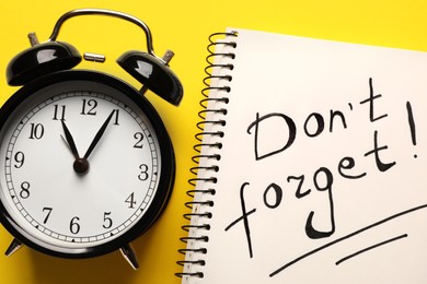 Photo of Alarm clock and notebook with phrase Don't forget! on yellow background, flat lay