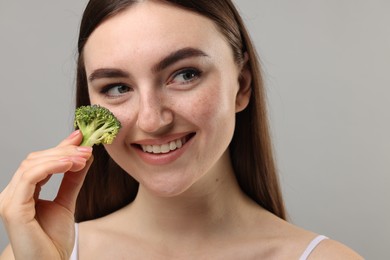 Photo of Smiling woman making fake freckles with broccoli and cosmetic product on grey background, closeup