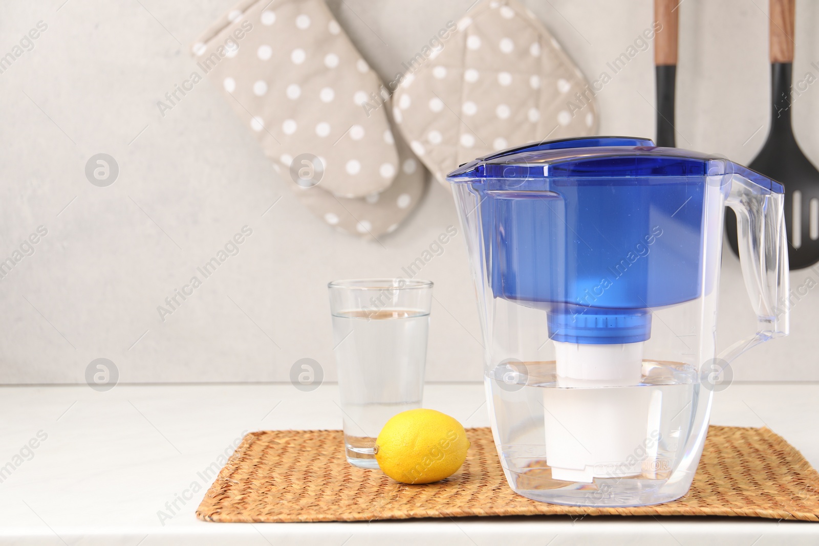 Photo of Water filter jug, glass and lemon on white countertop in kitchen, space for text