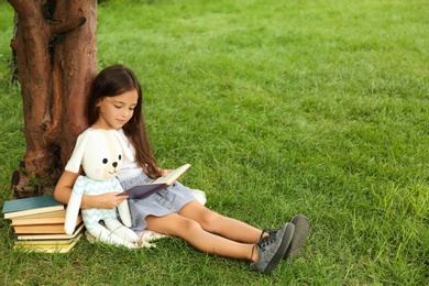 Cute little girl with toy reading book on green grass near tree in park
