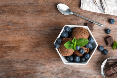 Flat lay composition with bowl of chocolate ice cream and blueberries on wooden table, space for text
