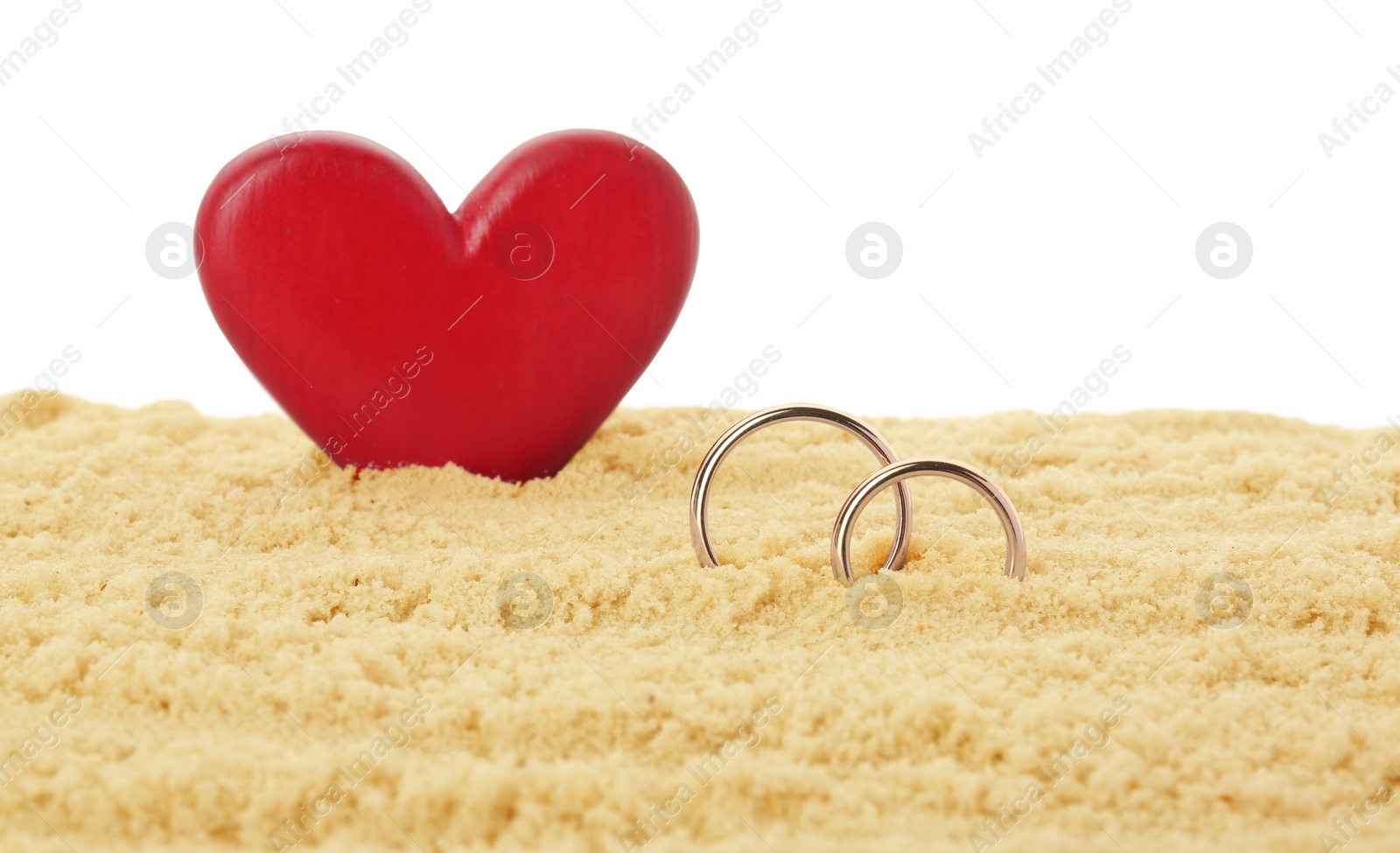 Photo of Honeymoon concept. Two golden rings, red wooden heart and sand isolated on white