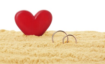 Photo of Honeymoon concept. Two golden rings, red wooden heart and sand isolated on white