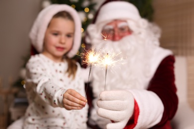 Photo of Santa Claus and little girl in room decorated for Christmas, focus on burning sparklers