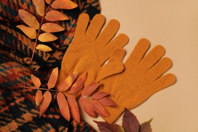 Stylish woolen gloves, scarf and dry leaves on beige background, flat lay