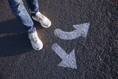 Choice of way. Man standing in front of drawn marks on road, closeup. White arrows pointing in different directions