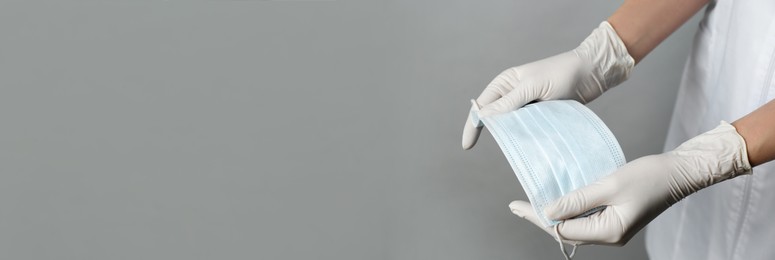 Doctor in medical gloves holding protective mask on light grey background, closeup. Banner design with space for text 