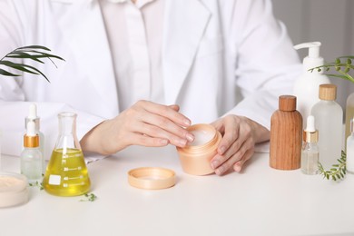 Photo of Dermatologist with jar testing cosmetic product at white table indoors, closeup