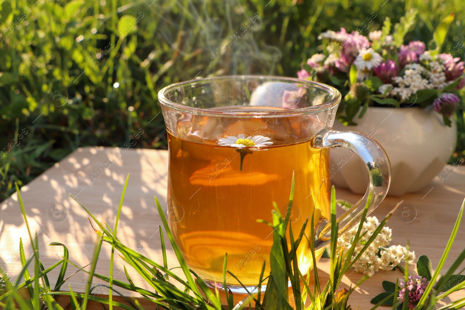 Photo of Cup of aromatic herbal tea and ceramic mortar with different wildflowers on green grass outdoors, closeup