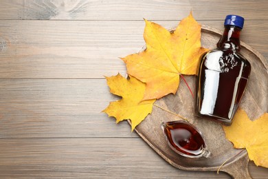 Photo of Bottle with tasty maple syrup, gravy boat and dry leaves on wooden table, top view. Space for text