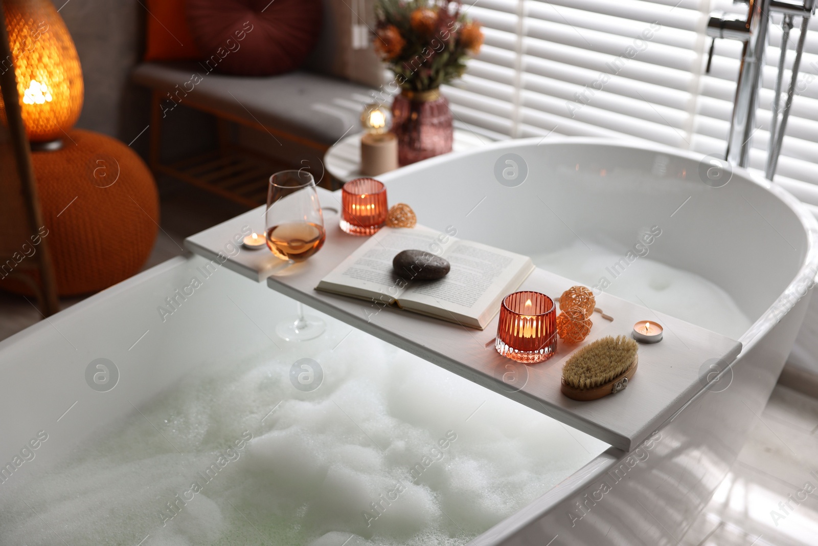 Photo of White wooden tray with glass of rose wine, book and burning candles on bathtub in bathroom