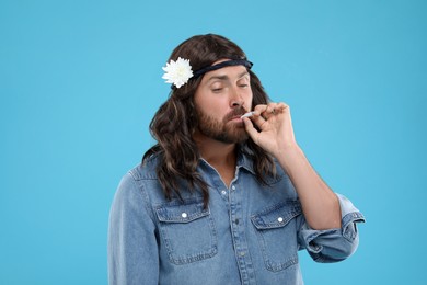 Stylish hippie man smoking cigarette on light blue background