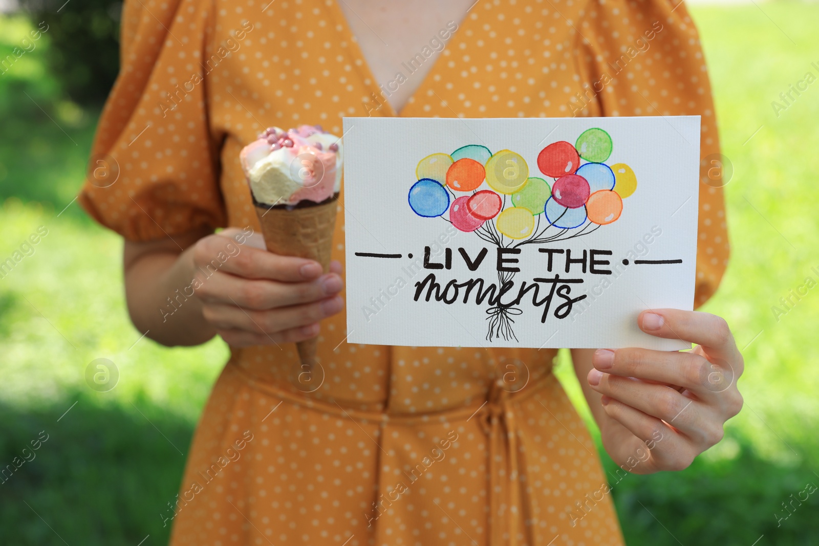 Photo of Woman holding card with phrase Live The Moments and ice cream outdoors, closeup
