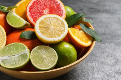 Photo of Different fresh citrus fruits and leaves in bowl on grey textured table, closeup