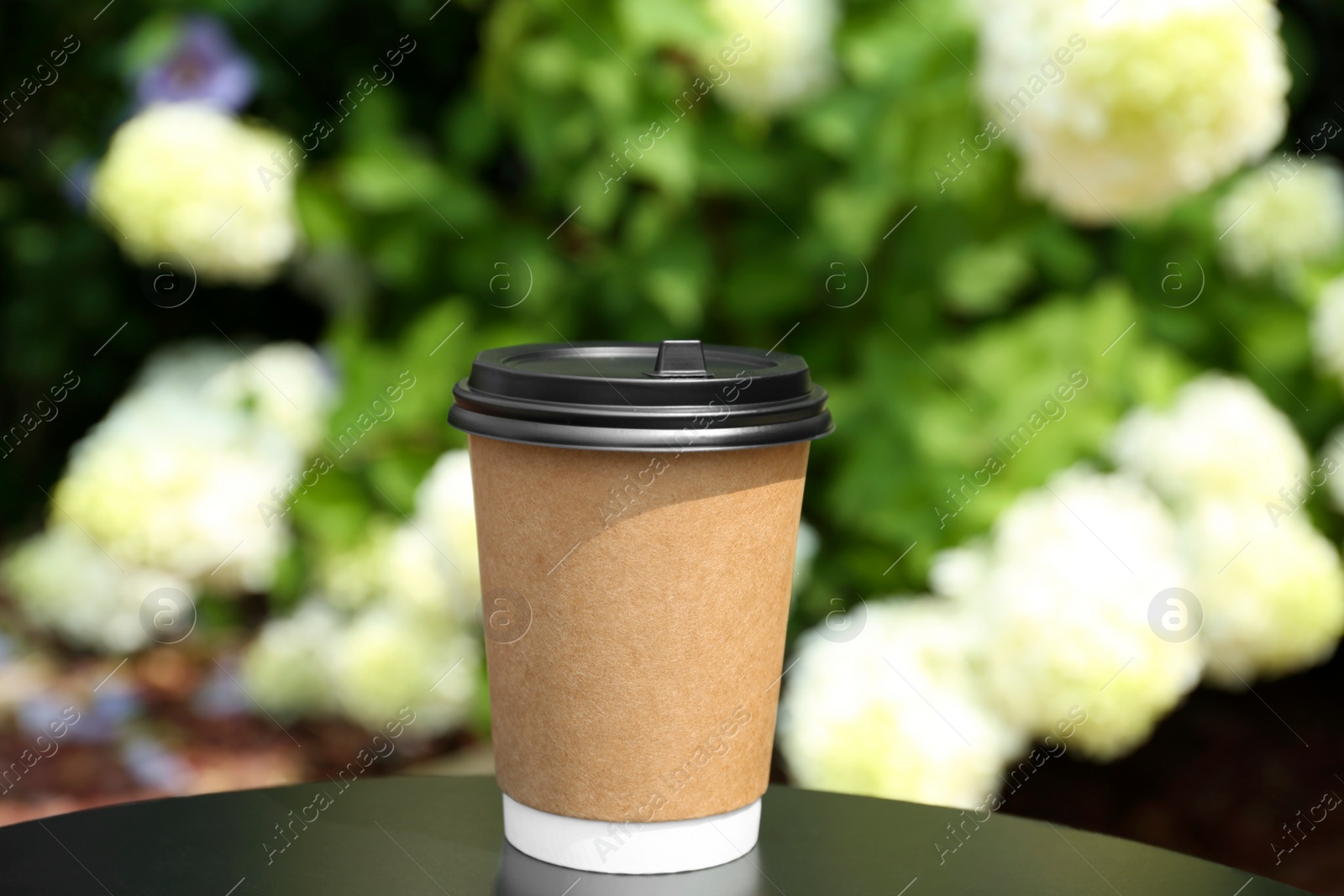 Photo of Cardboard cup with plastic lid on black table near flowers, space for text. Coffee to go