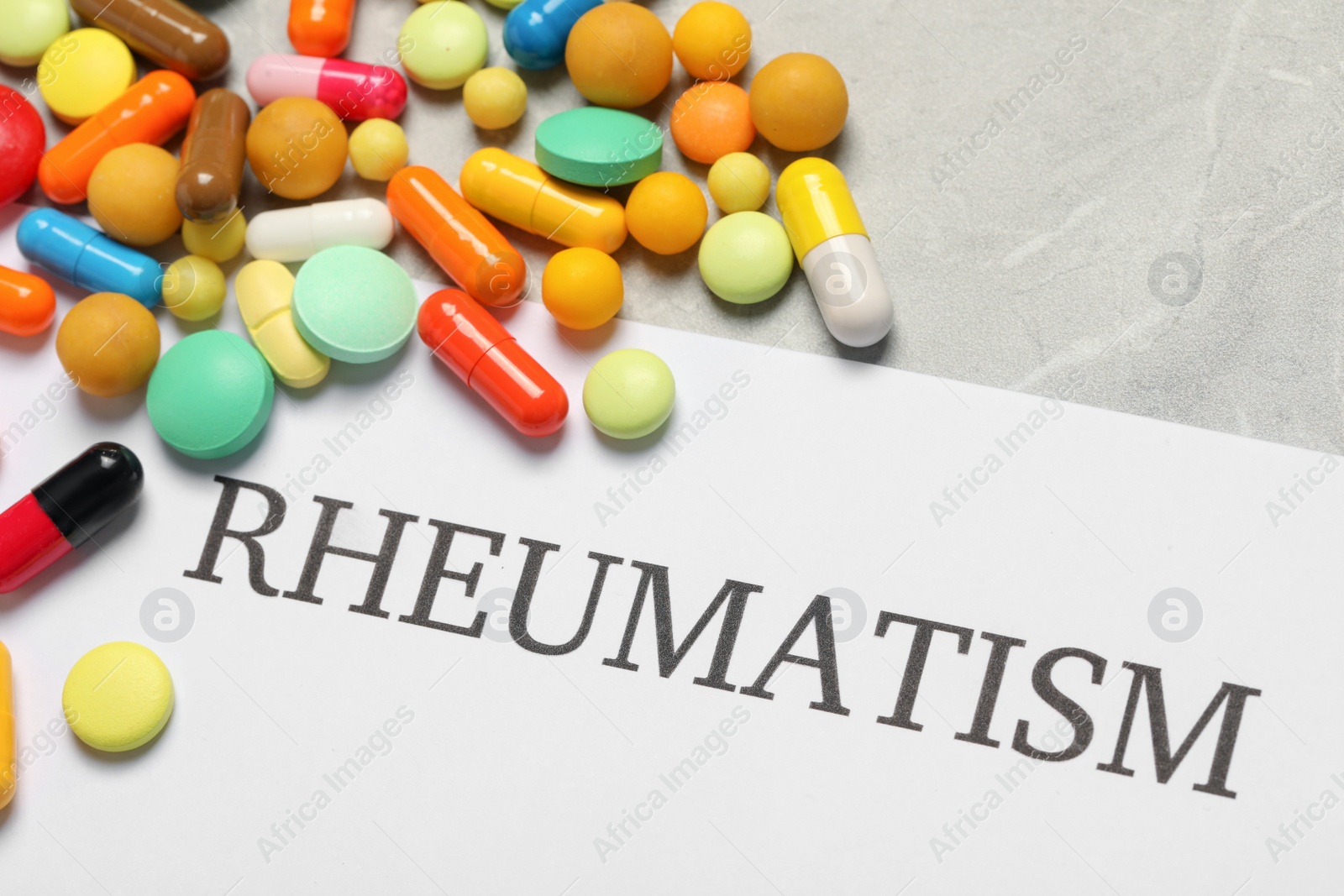 Photo of Paper with word Rheumatism and pills on light gray marble table, closeup