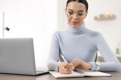 Photo of Young woman in glasses writing down notes during webinar at table in office