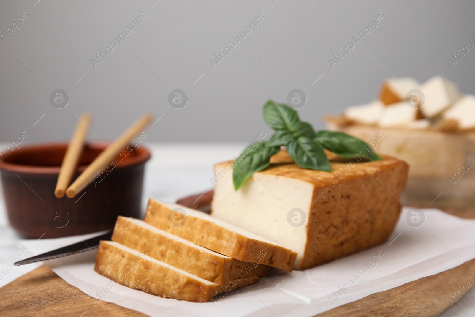 Photo of Board with smoked tofu, knife, basil and soy sauce on white wooden table