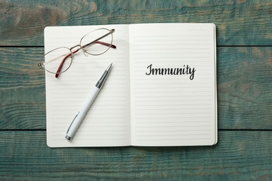 Photo of Notebook with word Immunity, glasses and pen on blue wooden table, top view