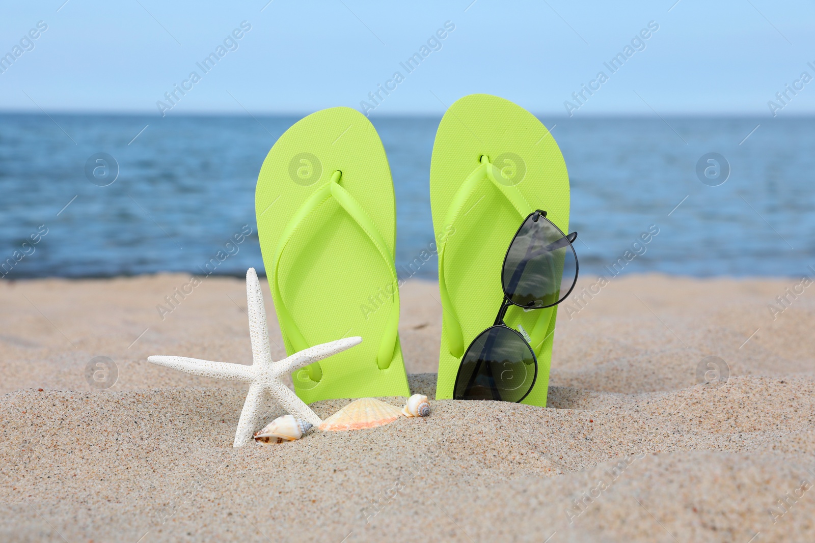 Photo of Stylish light green flip flops, sunglasses, starfish and seashells on beach sand