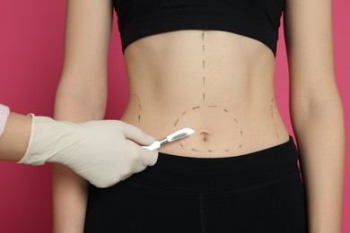 Photo of Doctor holding scalpel near slim woman with marks on body against pink background, closeup. Weight loss surgery