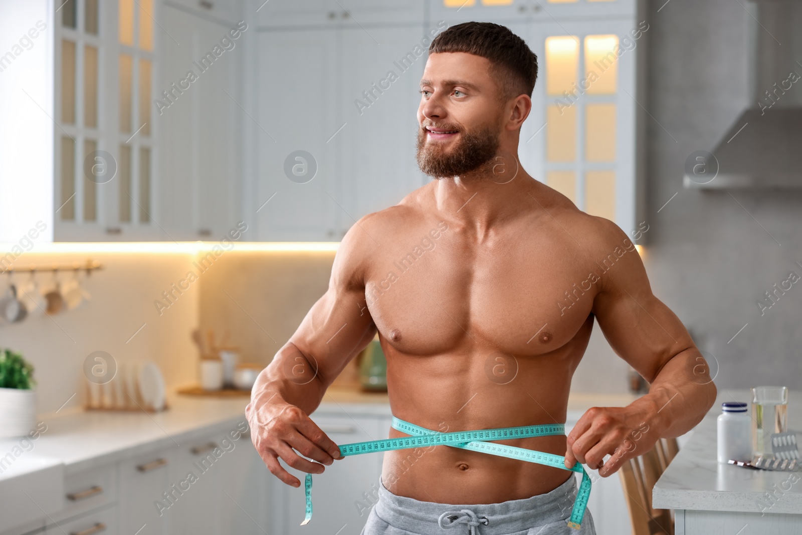Photo of Athletic young man measuring his waist with tape in kitchen, space for text. Weight loss