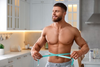 Athletic young man measuring his waist with tape in kitchen, space for text. Weight loss