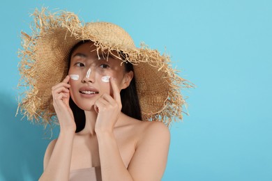 Photo of Beautiful young woman in straw hat with sun protection cream on her face against light blue background, space for text