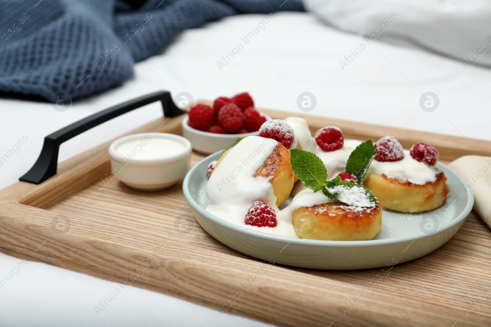 Photo of Tasty breakfast served in bedroom. Cottage cheese pancakes with fresh raspberries, mint, sour cream and icing sugar on wooden tray, closeup