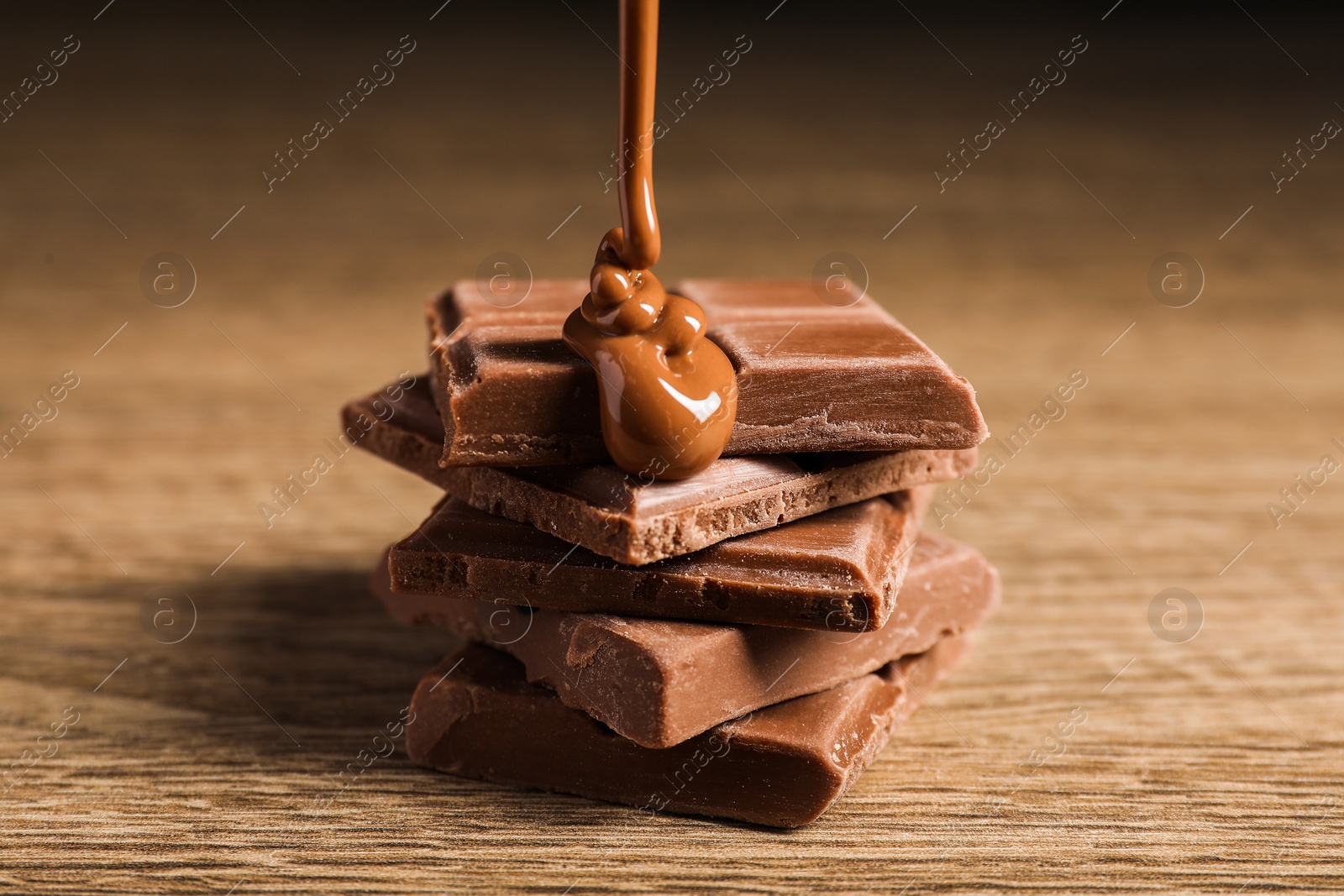 Photo of Pouring tasty milk chocolate paste onto pieces on wooden table, closeup