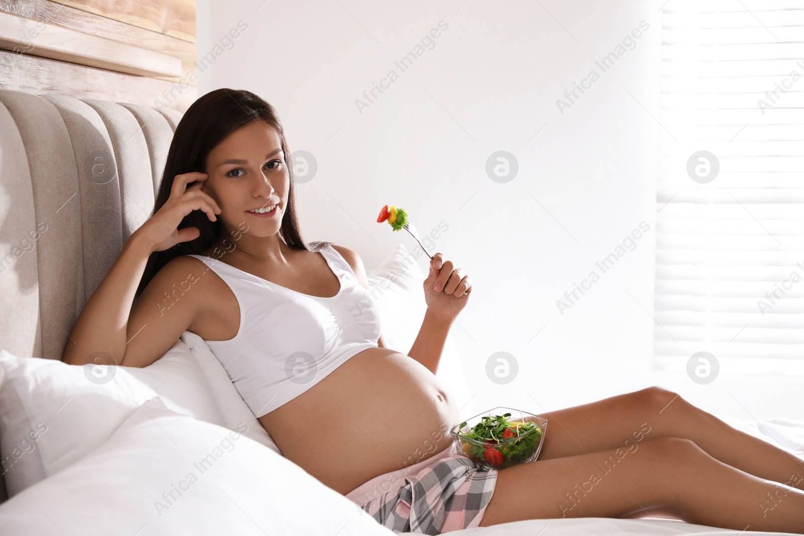 Photo of Young pregnant woman with bowl of vegetable salad in bedroom. Taking care of baby health