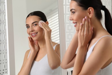 Photo of Beautiful woman removing makeup with cotton pad near mirror indoors