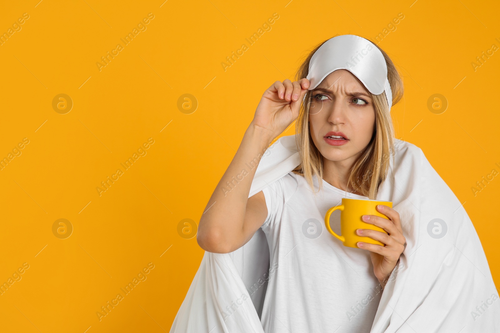 Photo of Young woman in sleeping mask wrapped with blanket holding cup on yellow background. Space for text