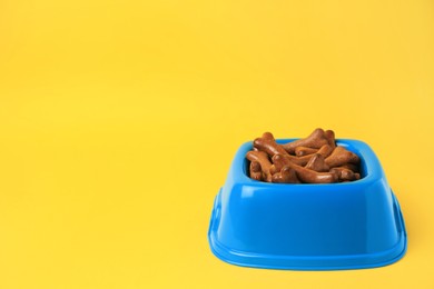Photo of Blue bowl with bone shaped dog cookies on yellow background, space for text