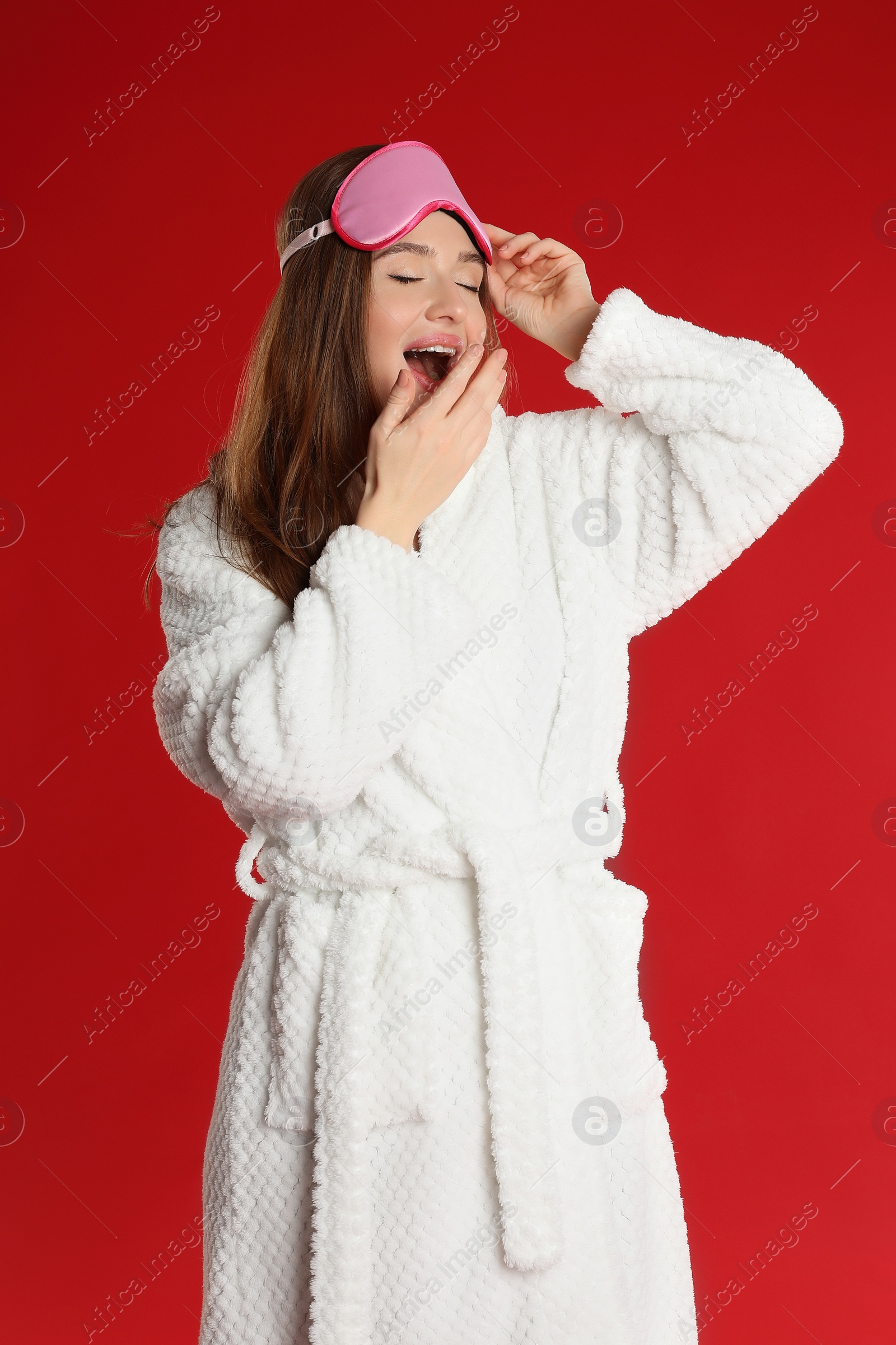 Photo of Young woman in bathrobe with eye sleeping mask yawning on red background
