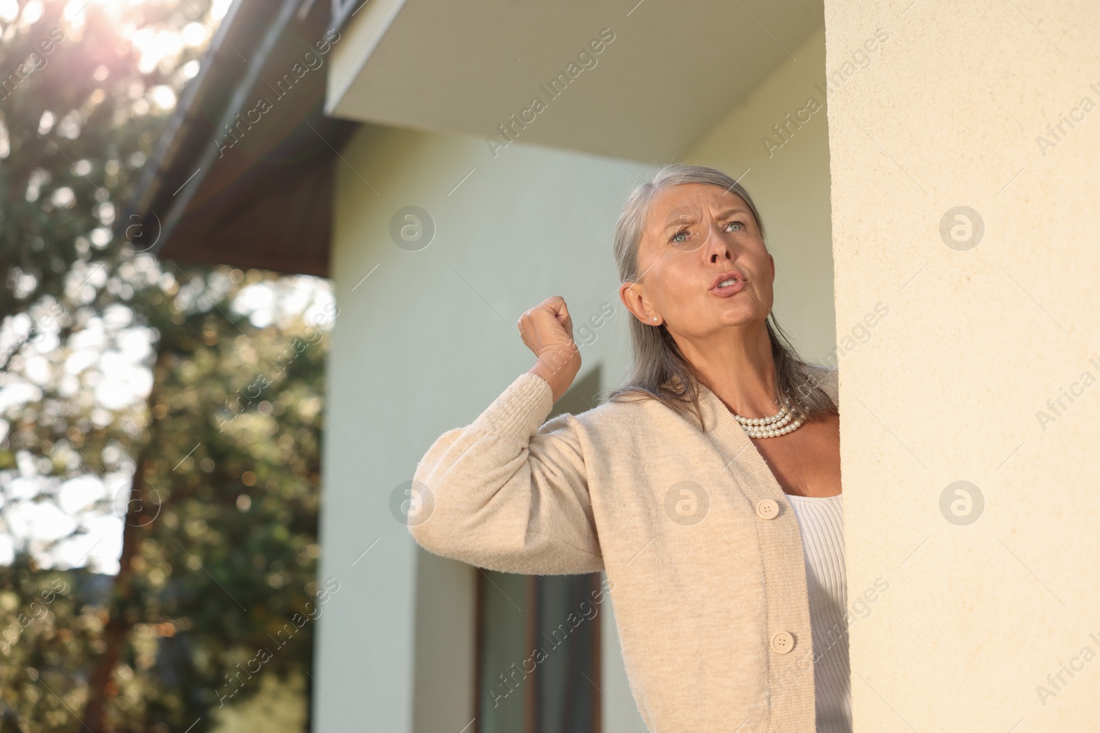 Photo of Angry senior woman showing fist near house, space for text. Annoying neighbour