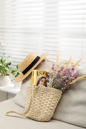 Stylish beach bag with wildflowers, magazine and book on sofa in room