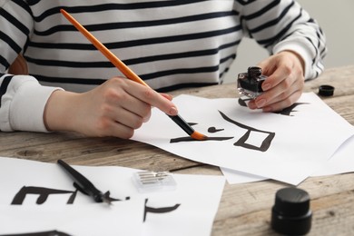 Calligraphy. Woman with brush and inkwell writing hieroglyphs on paper at wooden table, closeup