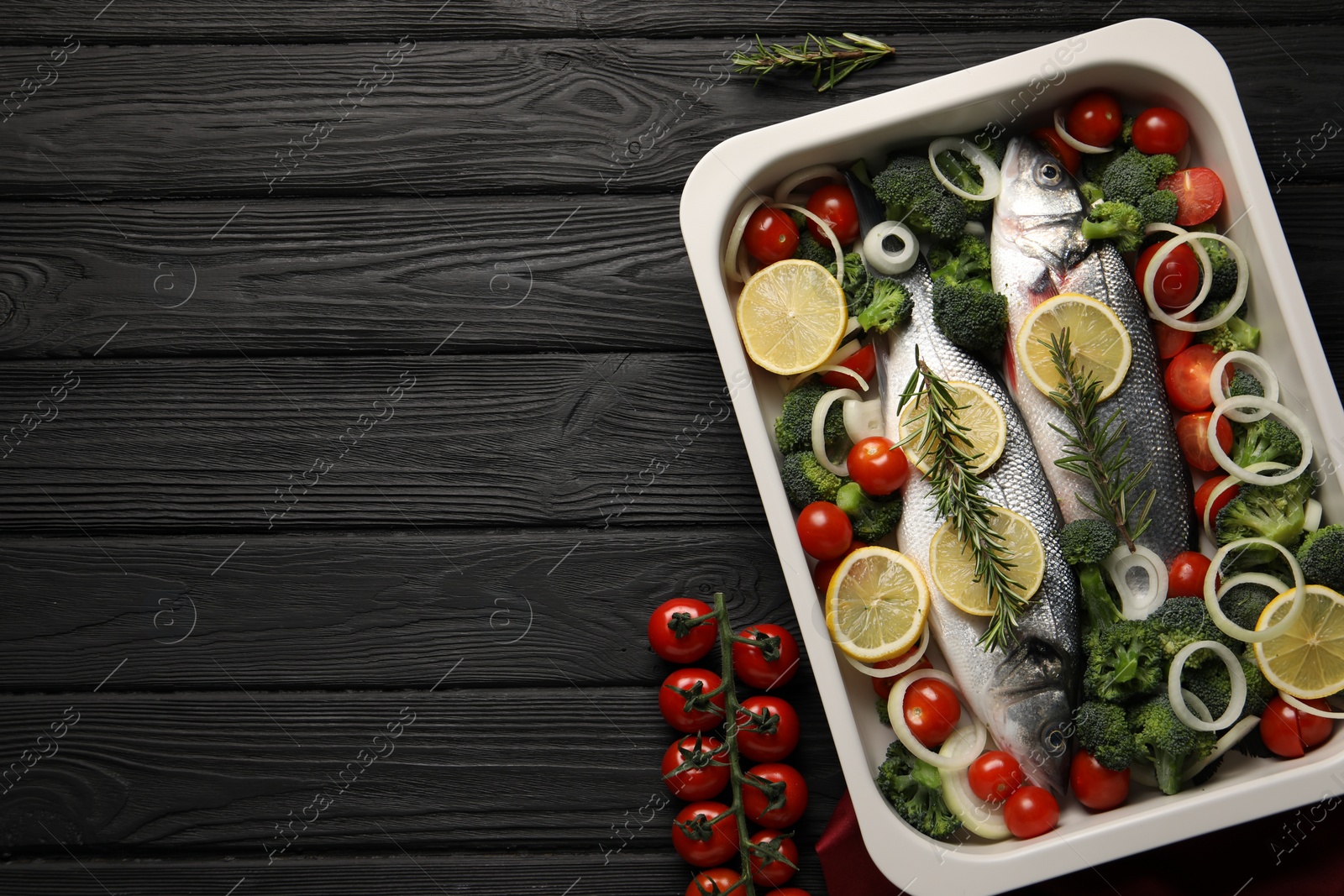 Photo of Raw fish with lemon in baking dish and vegetables on black wooden table, flat lay. Space for text