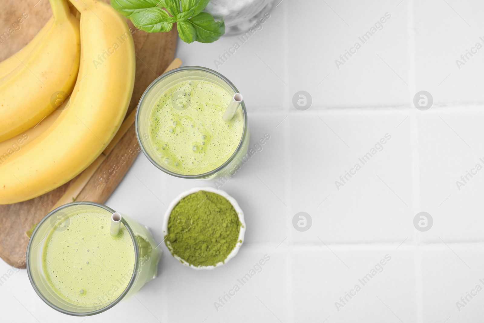 Photo of Glasses of tasty matcha smoothie, powder and bananas on white tiled table, flat lay. Space for text