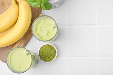 Photo of Glasses of tasty matcha smoothie, powder and bananas on white tiled table, flat lay. Space for text