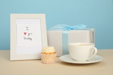 Photo of Happy Father's Day. Tasty cupcake, cup, card with phrase I Love You, Daddy in frame and gift box on beige table, closeup